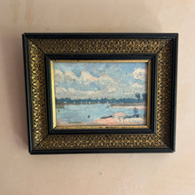 Carica l'immagine nel visualizzatore di Gallery, A framed landscape painting of a river scene with boats under a cloudy sky, enclosed in an ornate black frame with gold detailing, mounted on a beige wall.
