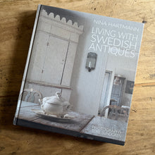 将图片加载到图库查看器，A hardcover book titled 'Living with Swedish Antiques' by Nina Hartmann, photographed on a rustic wooden tabletop. The cover features a muted, vintage photograph of a Swedish interior with a wooden cabinet and a teapot on a table.
